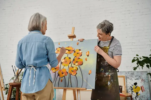 Two women stand in an art studio, one painting a canvas while the other admires the work. — Stock Photo