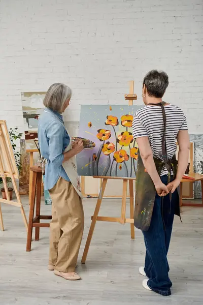 Two women in artists smocks, one painting orange flowers on canvas, the other observing closely in art studio — Stock Photo