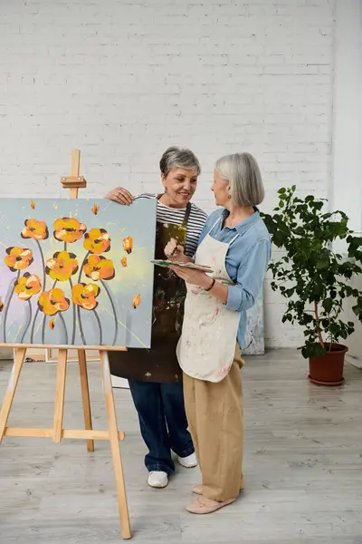 Due donne stanno in uno studio d'arte, una dipinge una tela su un cavalletto mentre l'altra tiene una tavolozza e osserva. — Foto stock