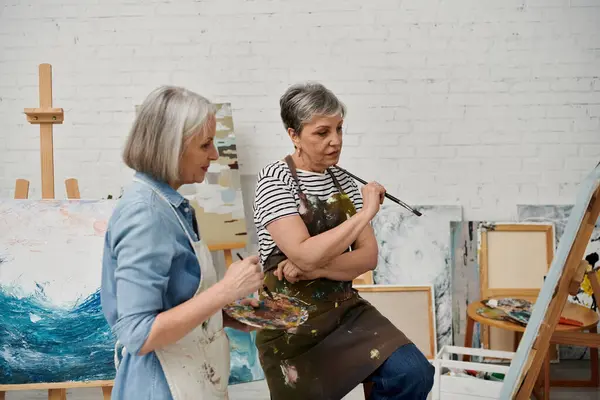 Deux femmes mûres, portant des blouses de peinture, se tiennent dans un atelier d'art, l'une tenant un pinceau, l'autre tenant une palette. — Photo de stock