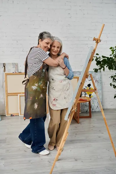 A loving lesbian couple in an art studio, embracing with paint-splattered aprons after a creative session. — Stock Photo