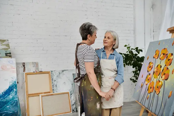 Two women in an art studio, dressed in painting aprons, hold hands and look at each other lovingly. — Stock Photo