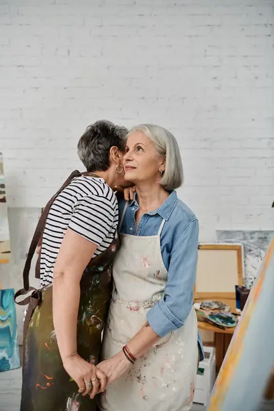A lesbian couple, wearing paint-splattered aprons, share a moment in their art studio. — Stock Photo