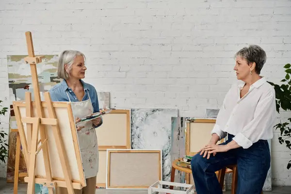 Two mature women in an art studio, one holds a paint palette while the other sits and observes. — Stock Photo