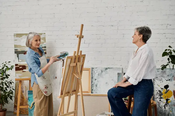 Two mature women paint together in an art studio, one holding a paintbrush and palette while the other watches. — Stock Photo