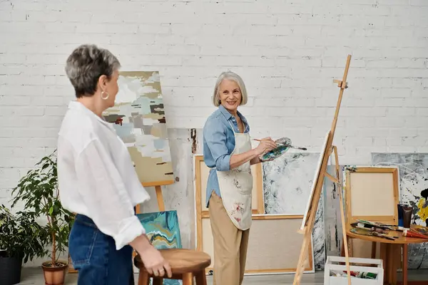Two mature women are in an art studio. One woman is painting on a canvas while the other woman watches. — Stock Photo