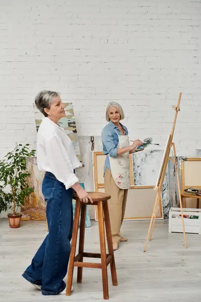 Deux femmes dans un studio d'art. L'un peint sur une toile tandis que l'autre se penche contre un tabouret, regardant. — Photo de stock