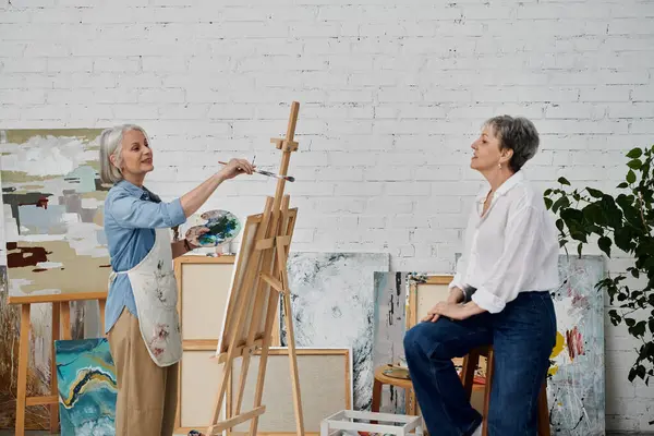 A mature lesbian couple paints together in a light and airy studio, one woman working on a canvas while the other observes. — Stock Photo