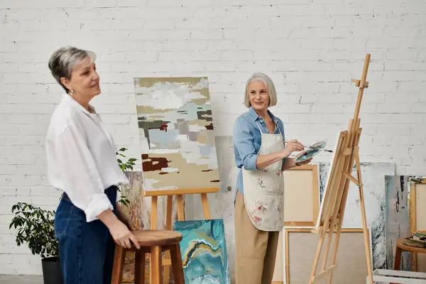 Two mature lesbian women in an art studio, one painting while the other posing. — Stock Photo