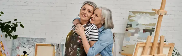 A couple in an art studio, one woman paints while the other lovingly embraces her from behind. — Stock Photo