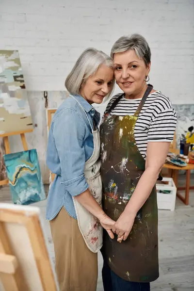 Two older women, a lesbian couple, hold hands in a painting studio, surrounded by art supplies and canvases. — Stock Photo