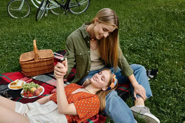 Dos mujeres disfrutan de una cita de picnic en un parque verde, con una mujer acostada en la otra vuelta. - foto de stock