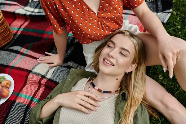 A young woman lays on a checkered blanket in a park, her girlfriend resting her hand on her. — Stock Photo