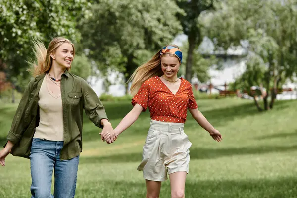 Two young women, holding hands, walk through a lush green park on a sunny day. — Stock Photo
