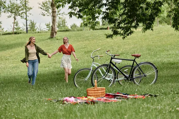 Two young women, dressed in casual clothing, hold hands and walk away from their bicycles in a green park. — Stock Photo