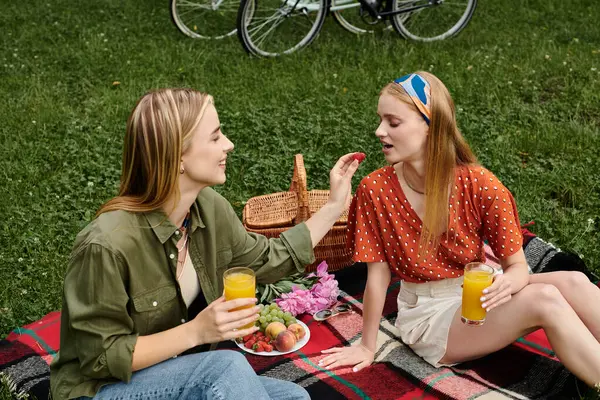 Duas jovens mulheres desfrutando de um piquenique juntas em um parque verde, compartilhando um lanche e bebidas. — Fotografia de Stock