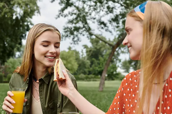 A young lesbian couple shares a sandwich and a smile on a sunny afternoon in the park. — Stock Photo
