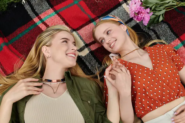 Two young women lie on a blanket in a park, holding hands and smiling at each other. — Stock Photo