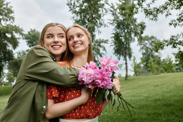 Two women embrace in a verdant park, a bouquet of pink peonies adding a touch of romance to the scene. — Stock Photo