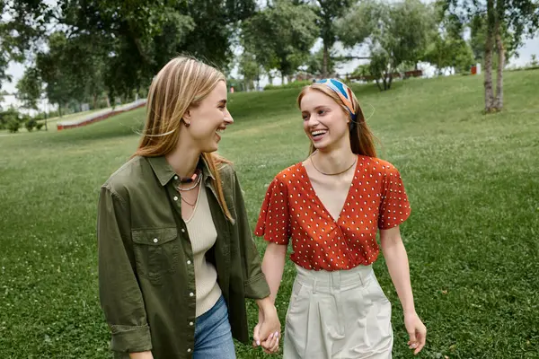 A young lesbian couple laughs as they walk hand-in-hand through a grassy park. — Stock Photo