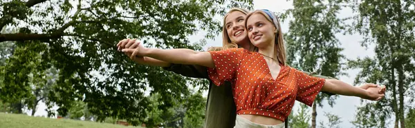 A young lesbian couple enjoys a playful moment in a green park, their arms outstretched and faces lit with happiness. — Stock Photo
