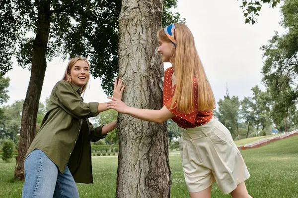 Two young women share a laugh while leaning against a tree in a park. — Stock Photo