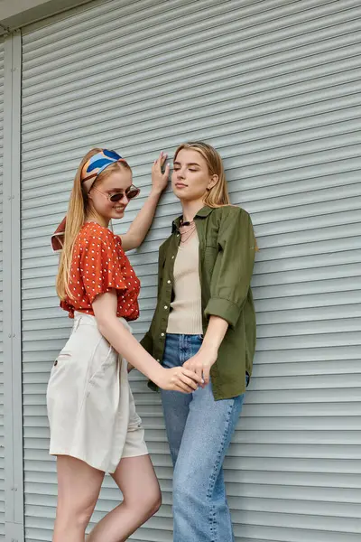 Dos mujeres jóvenes, vestidas con atuendo casual, se toman de la mano y sonríen apoyadas en una pared - foto de stock