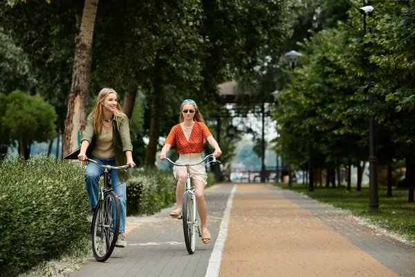 Two young women ride bicycles side-by-side on a paved path through a lush green park. — Stock Photo