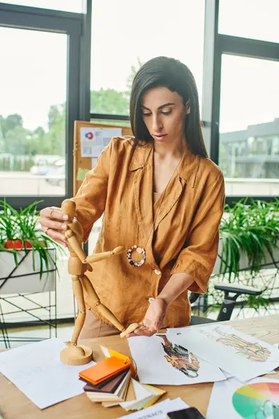 A woman in a tan blouse adjusts a wooden fashion doll while working on sketches at a desk in an office. — Stock Photo