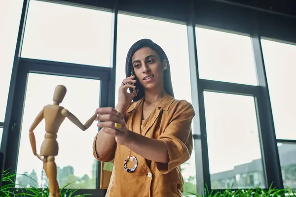 A woman in a bright yellow shirt on a phone call with a wooden doll in front of her. — Stock Photo