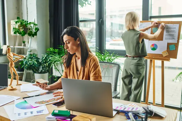 Una mujer de Oriente Medio con el pelo largo y oscuro trabaja en su escritorio en una oficina moderna, rodeada de elementos de diseño coloridos y un colega - foto de stock