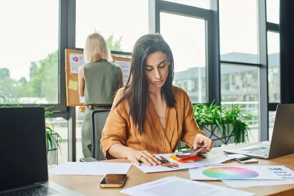 Una mujer en una blusa de color amarillo brillante examina una paleta de colores, perdida en el pensamiento mientras trabaja en un entorno de oficina contemporáneo. - foto de stock