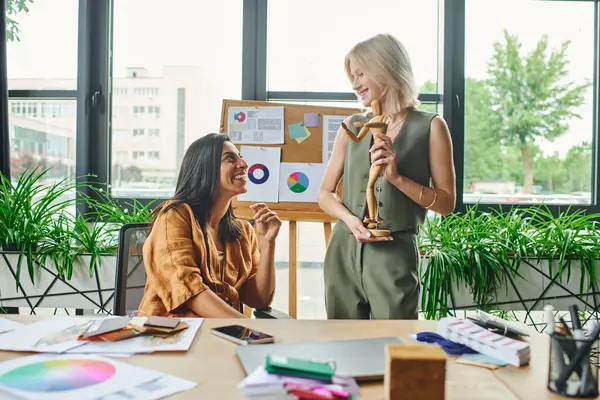 Two women discuss design ideas in a modern office, one holding a wooden figurine. — Stock Photo