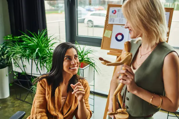 Two women discussing a design concept: a brunette in a brown blouse looking up at her blonde coworker holding a wooden figurine — Stock Photo
