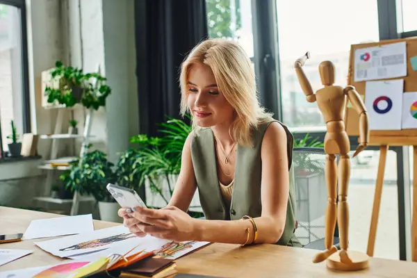 Una donna bionda in un gilet elegante lavora alla sua scrivania, controllando il telefono e perso nel pensiero, con un manichino di legno in piedi nelle vicinanze. — Foto stock