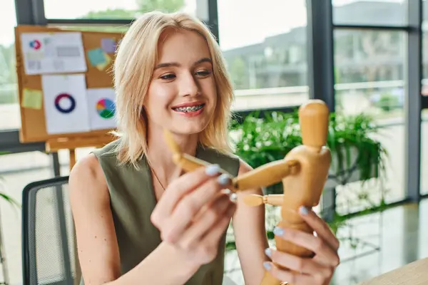 A blonde woman in a green tank top smiles while posing a wooden figure in an office setting. — Stock Photo