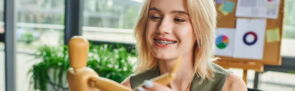 A young blonde woman in a casual office outfit smiles as she works with a wooden figure, surrounded by office supplies and natural light. — Stock Photo