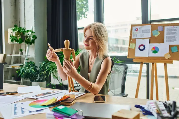 Une femme blonde dans une tenue décontractée intelligente travaille dans un bureau, tenant un mannequin en bois et étudiant des plans de design. — Photo de stock