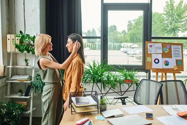Two women stand in a bright, modern office, their heads tilted towards each other in a shared moment of connection. — Stock Photo