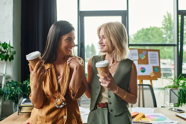 Two women in smart casual attire, working in a modern agency, enjoy a coffee break, exchanging ideas and smiles. — Stock Photo