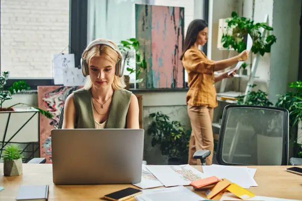 Deux femmes en tenue de travail travaillent dans un bureau moderne - l'une sur un ordinateur portable, l'autre examinant des documents en arrière-plan — Photo de stock