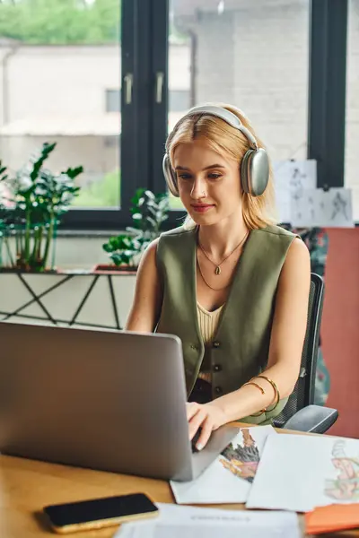 Eine junge Frau, die Kopfhörer und schicke Freizeitkleidung trägt, arbeitet in einem gemütlichen Büroambiente emsig an einem Laptop.. — Stockfoto