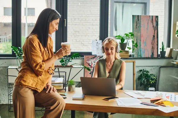 Two young women work together on a project in a modern agency, one holds a coffee cup while the other focuses on her laptop. — Stock Photo