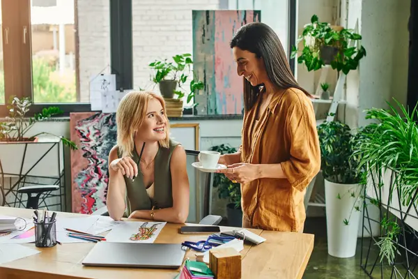 Two women in smart casual attire work together on a project in a modern office space. One woman offers a cup of coffee to her partner. — Stock Photo