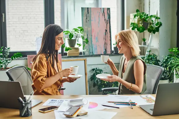 Two women, dressed in smart casual attire, enjoy a coffee break while working on a project in a contemporary office setting. — Stock Photo