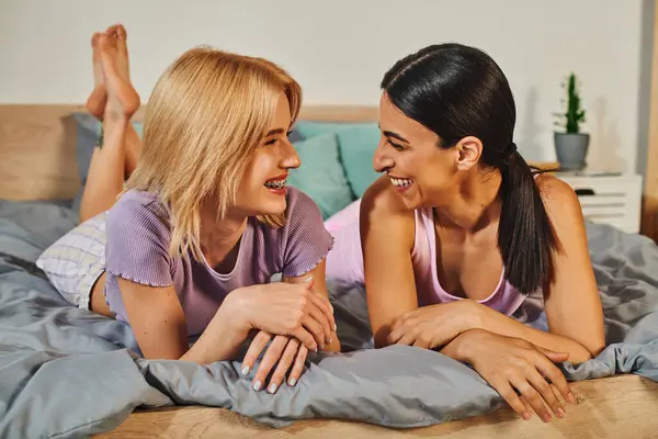 Dos mujeres, una con el pelo rubio y la otra con el pelo oscuro, se acuestan juntas en una cama, sonriendo y abrazándose. - foto de stock