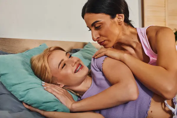 Deux femmes, une blonde et une brune, se blottissent sur un lit dans une maison. La blonde se couche sur un oreiller tandis que la brune la serre dans ses bras, les deux souriantes — Photo de stock