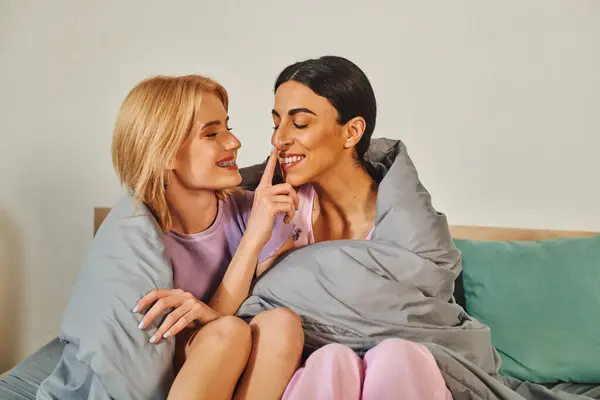 Duas mulheres se abraçam debaixo de um cobertor em sua cama, compartilhando um momento lúdico. — Fotografia de Stock