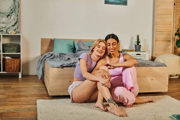 Un couple de lesbiennes, l'une aux cheveux blonds et l'autre aux cheveux foncés, s'assoient sur un tapis dans leur maison, les bras enroulés l'un autour de l'autre. — Photo de stock