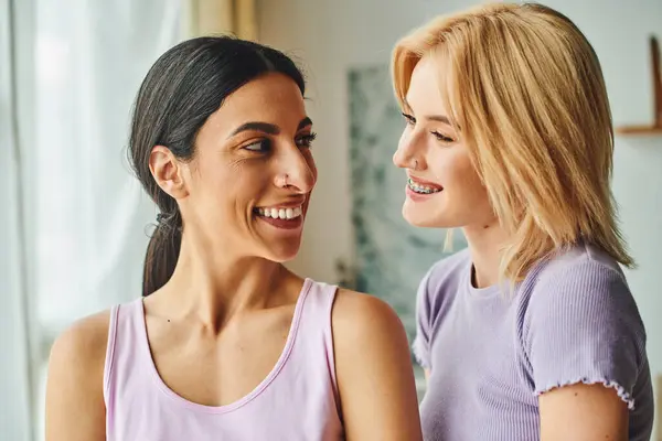 Two women, one with long black hair and the other with blonde hair, smile at each other in a cozy home setting. — Stock Photo